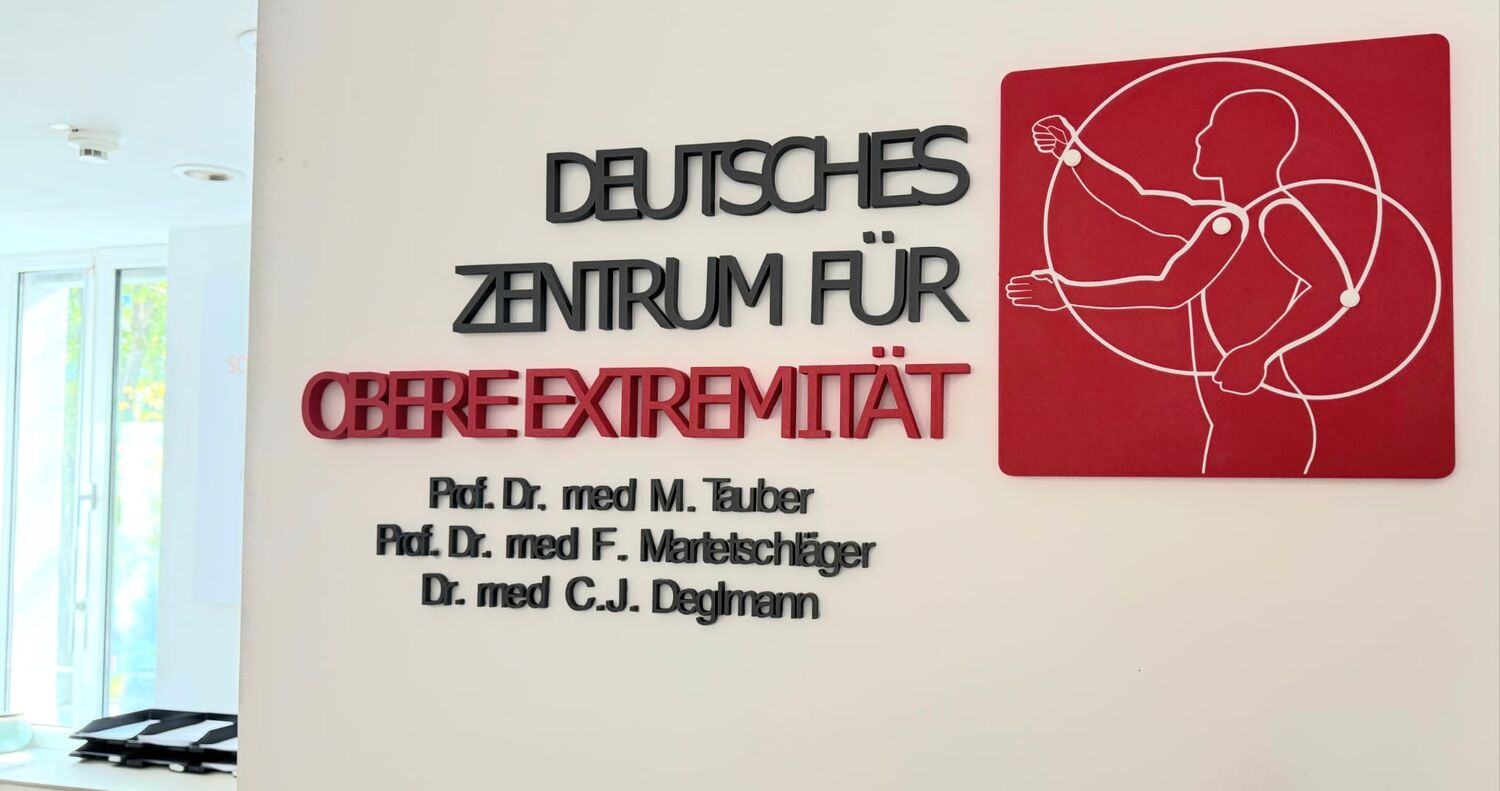 3D-Wandlogo des Deutschen Zentrums für obere Extremität mit schwarzer und roter 3D-Schrift und roter Logotafel.