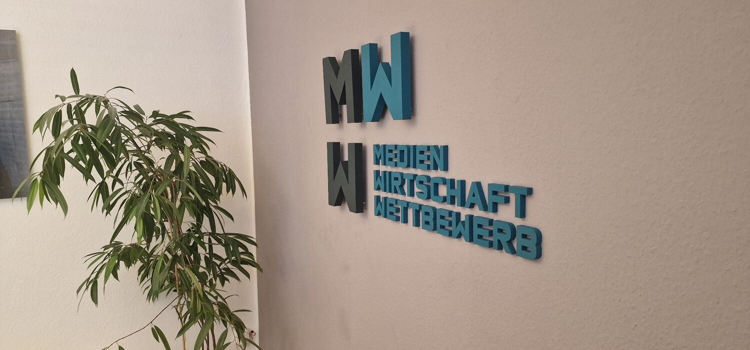 3D-Wandlogo „Medien Wirtschaft Wettbewerb“ in Schwarz und Blau an einer Bürowand.